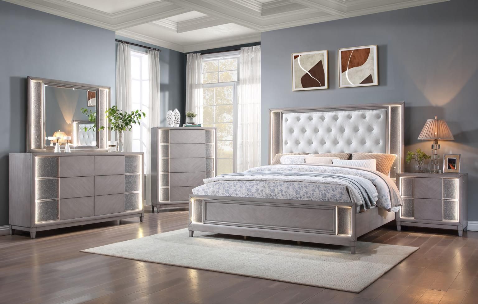 Light grey wood bedroom set with silver accents and led lighting. Headboard has upholstered center with tufting. there is a dresser, mirror, nightstand and 5-drawer chest.