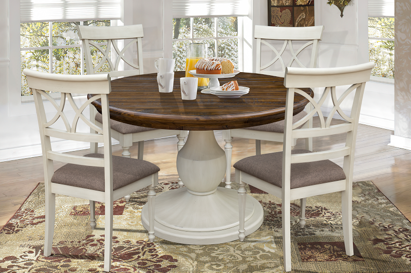 The Brockton Dining Set features a solid wood round table with a pedestal base, finished in wheat color and a dark brown top, both with a distressed look. It includes four side chairs, each with upholstered seat cushions.