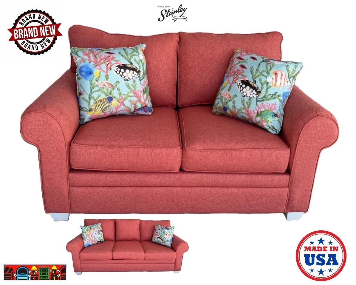 A sofa in coral color or an exact copy in a queen-sized sleeper. It has rolled arms, three cushions for seating, and two throw pillows with a coastal print that contrast with the sofa color. The sofa rests on white feet.