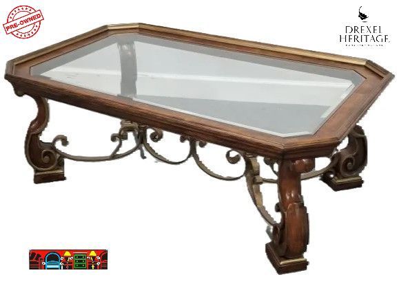 'A pre-owned cocktail table by Drexel Heritage, crafted from solid wood in a medium brown hue, featuring elegant gold accents and decorative iron elements on the base, complemented by an inset glass top.'