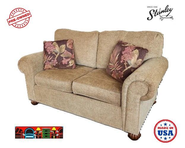 This gently used loveseat has two seats and rolled arms in a golden-brown color. It comes with two throw pillows that have a tropical leaf print in contrasting colors. The cushions are reversible, and the feet are dark brown and round.
