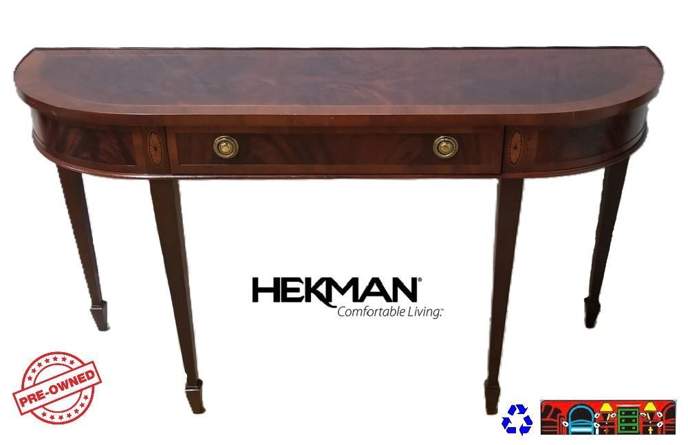 The gently used Hekman Sofa Table is crafted from Mahogany solids, featuring a Yae wood border atop with a swirl mahogany inset on both the top surface and the drawer front. This half-moon-shaped table stands on four slender legs, accented with Ebony inlay. It includes one drawer, adorned with Brass hardware and finished in the sophisticated Copley Place style.