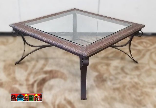A gently used square cocktail table featuring a brown iron base, a top with faux painting, and an inlay of beveled glass.