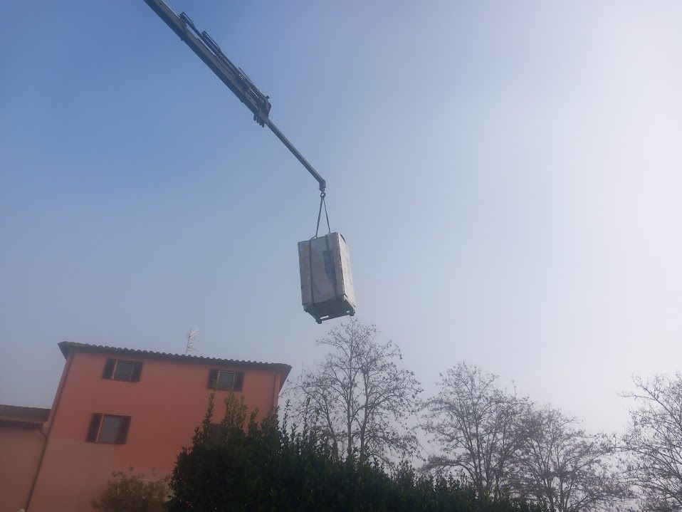 Una gru solleva un grande container bianco sopra un edificio rosso e alberi sotto un cielo azzurro.