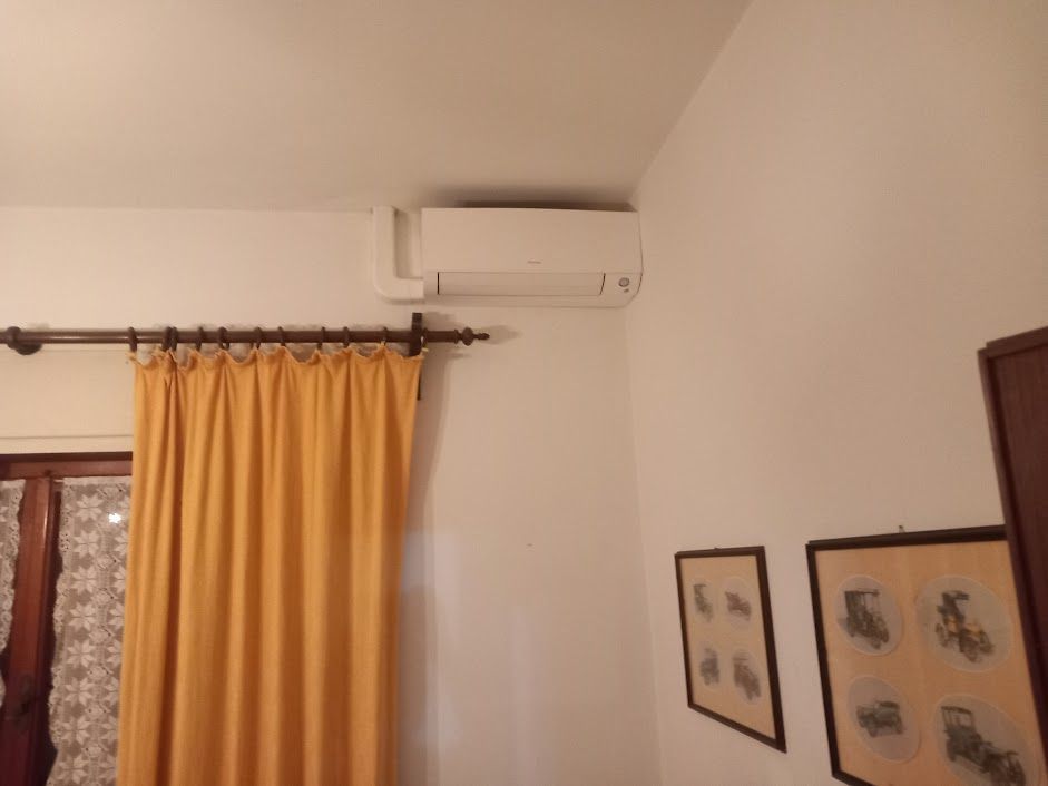 Un condizionatore d'aria è montato su una parete bianca sopra una finestra con tende gialle. Sotto sono appese due foto incorniciate.