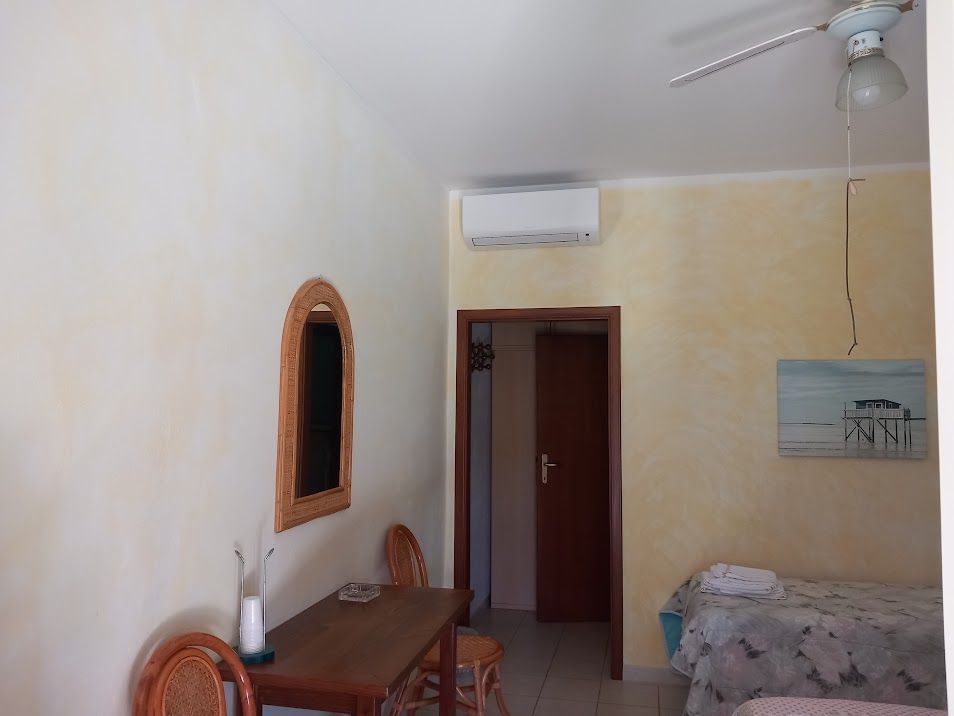 Interno di una stanza con due letti, un tavolo e uno specchio. Pareti chiare, mobili in legno chiaro e una porta che conduce a un'altra stanza.