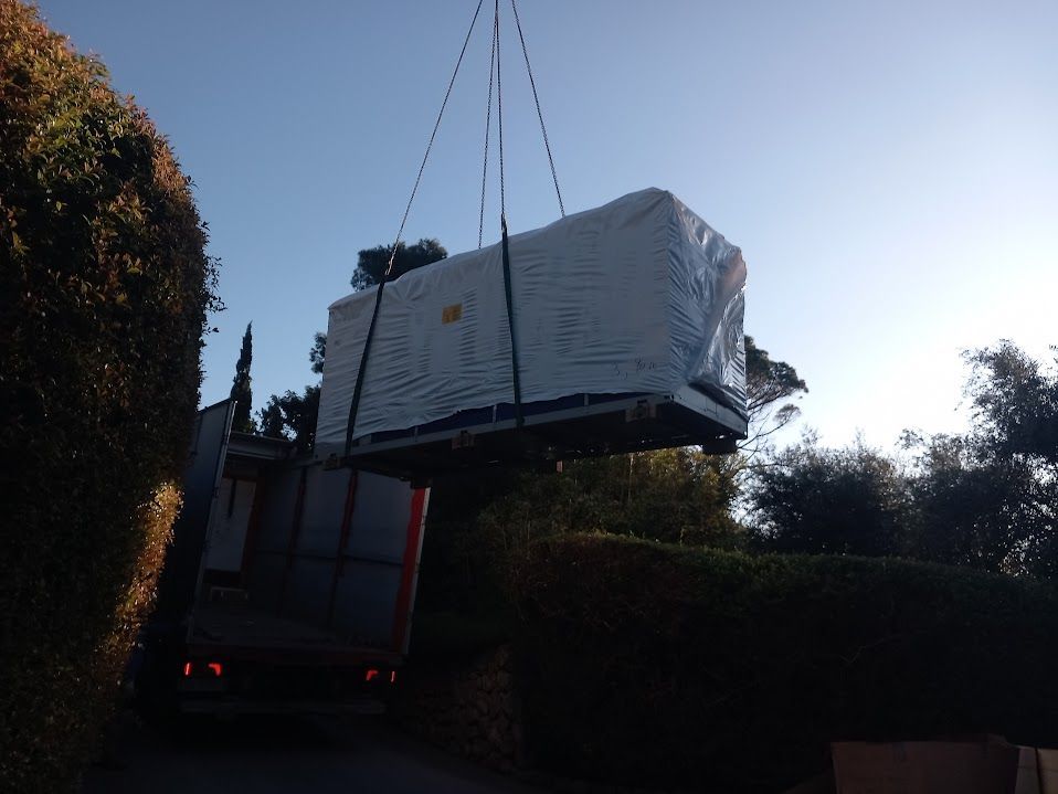 Un grande oggetto avvolto, sospeso da una gru sopra un camion in un vialetto fiancheggiato da cespugli verdi.