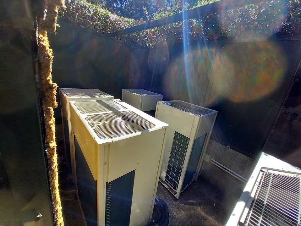 Unità HVAC all'interno di un'area chiusa. Macchine beige e grigie con ventole. Luce solare intensa.