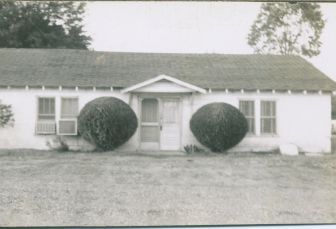 Black and white photo of a white, single-story house with two large bushes flanking the front door.