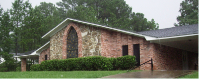 Brick building with arched window and dark roof, set against green trees.