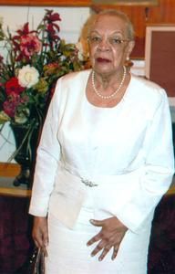 Woman in white suit, standing indoors, near flowers. Wearing glasses and pearl necklace.