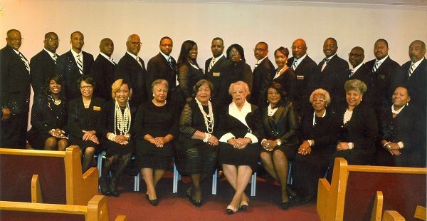 Group of people, formal attire, in a church. Men in suits stand behind women seated in front.