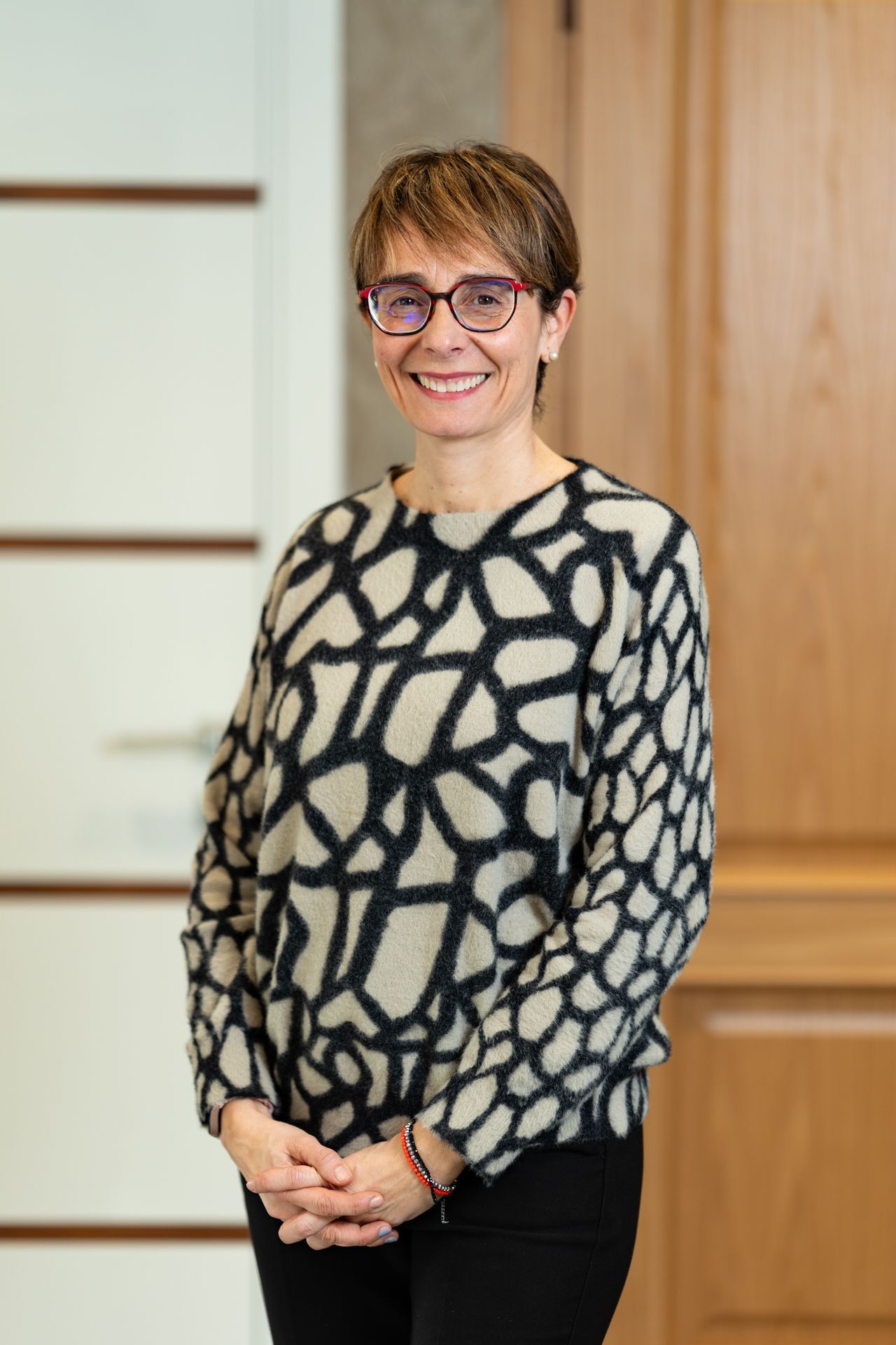Una donna che indossa un maglione fantasia e occhiali sorride, in piedi vicino a porte di legno.