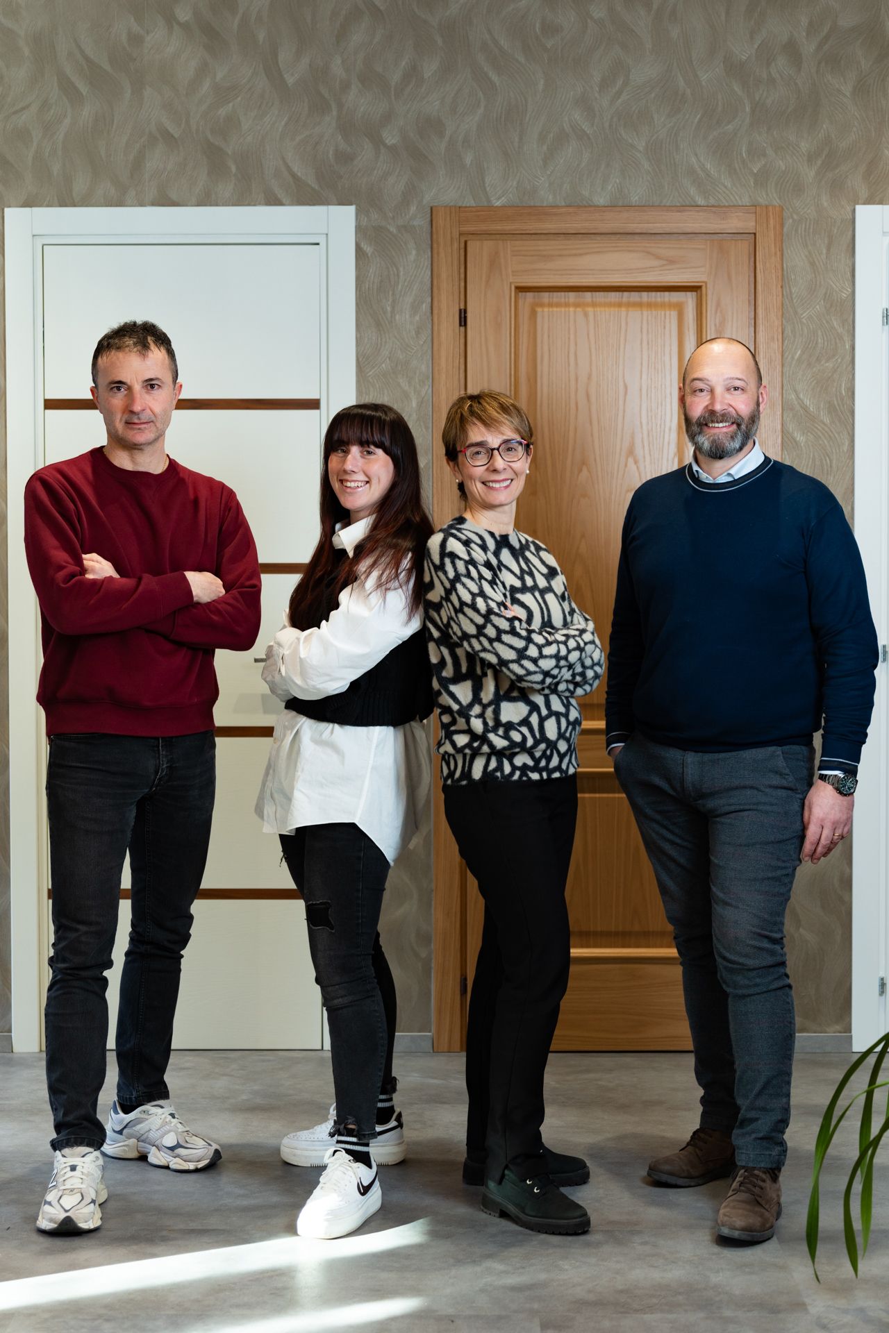 Quattro persone in piedi davanti a una porta, con le braccia incrociate. Due uomini e due donne, in abiti casual. Ambientazione da studio.