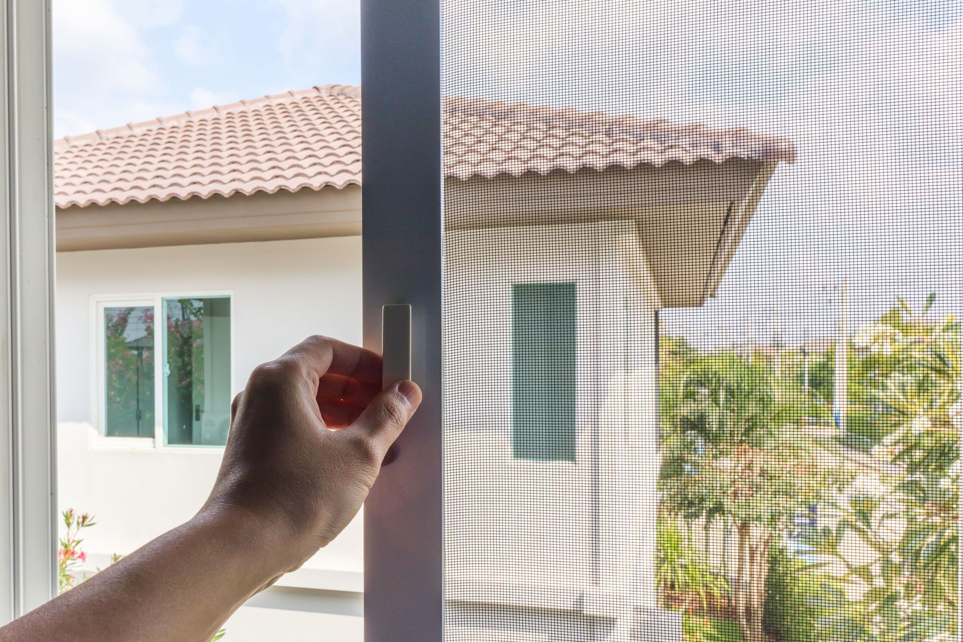 Mano che apre una zanzariera, con una casa e del verde visibili all'esterno.