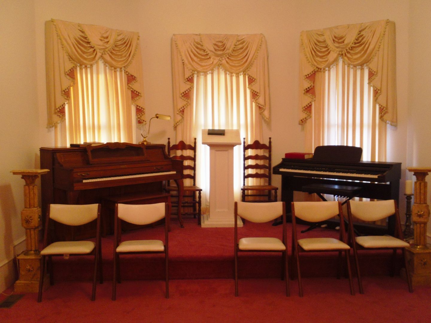 Interior of a room with a red carpet, stage with two pianos, chairs, and podium, and three windows with curtains.