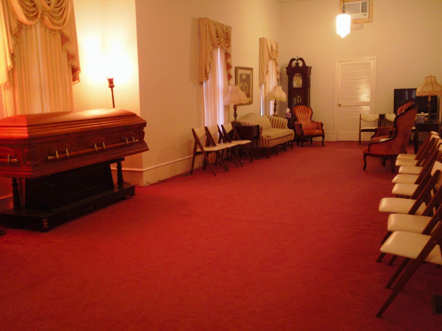 A casket in a funeral home; red carpet, chairs, clock, and seating area.