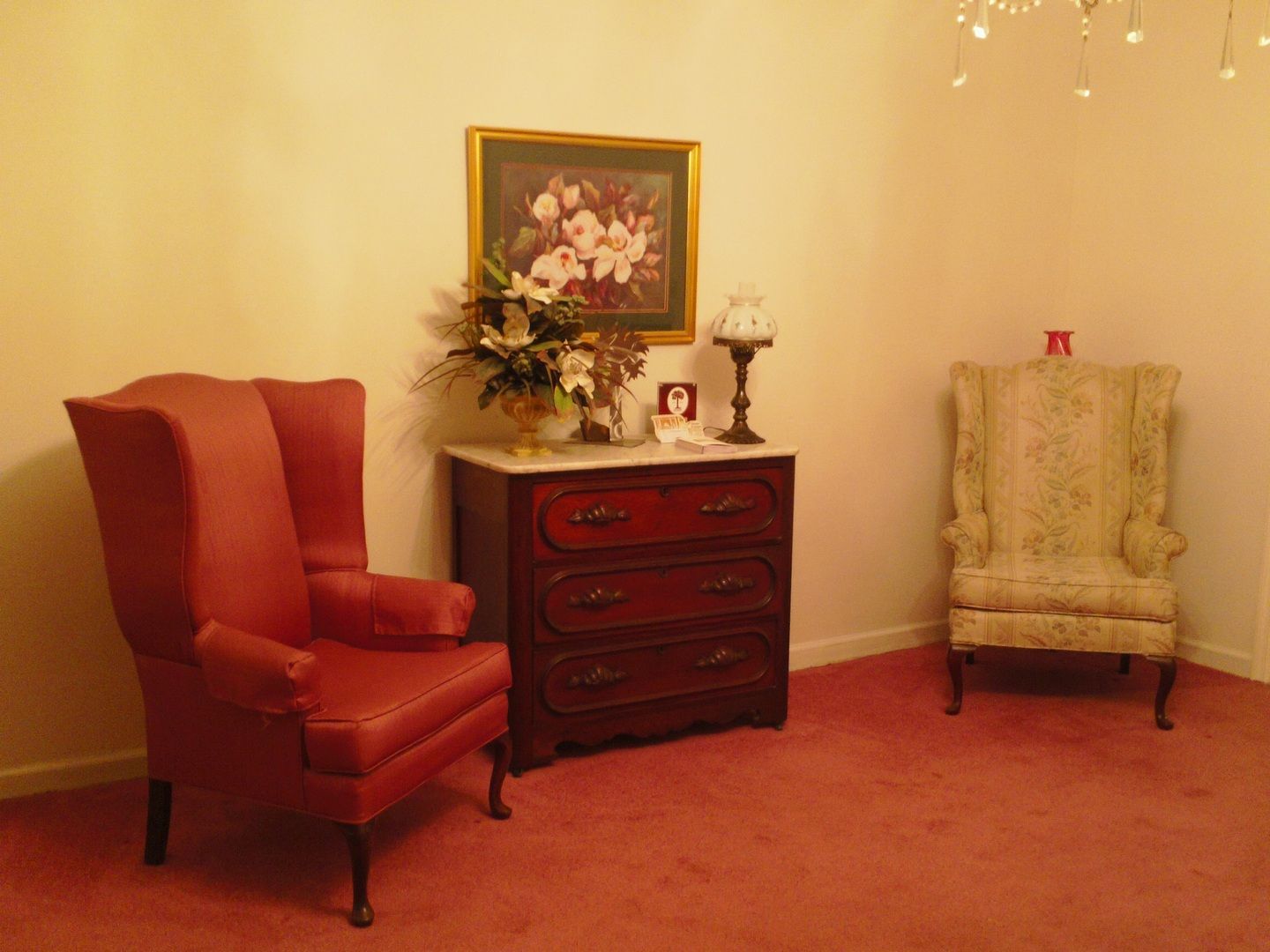 Red upholstered chair and vintage patterned armchair, chest with floral arrangement and picture, under a framed painting, on red carpet.