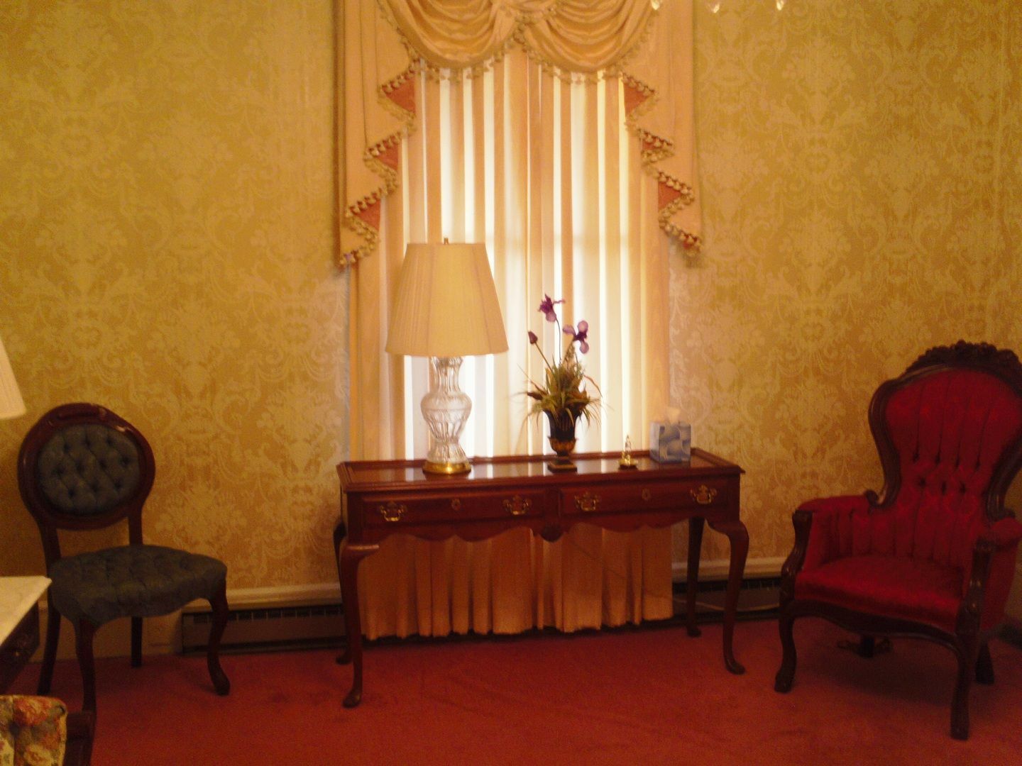 A formal room with a red carpet, two chairs, a table with a lamp, and a window with curtains.