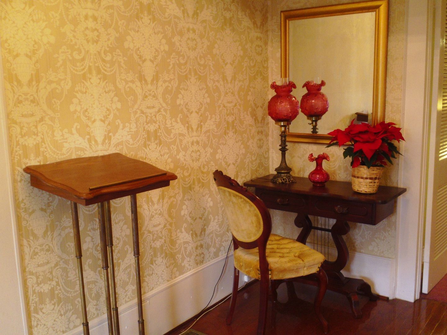 Corner with a dark table, mirror, and two red lamps. A chair and a plant. Light floral wallpaper.