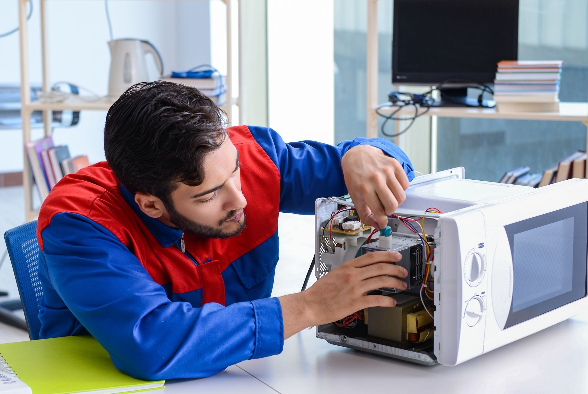 A person in work clothes repairing a white microwave in an office.