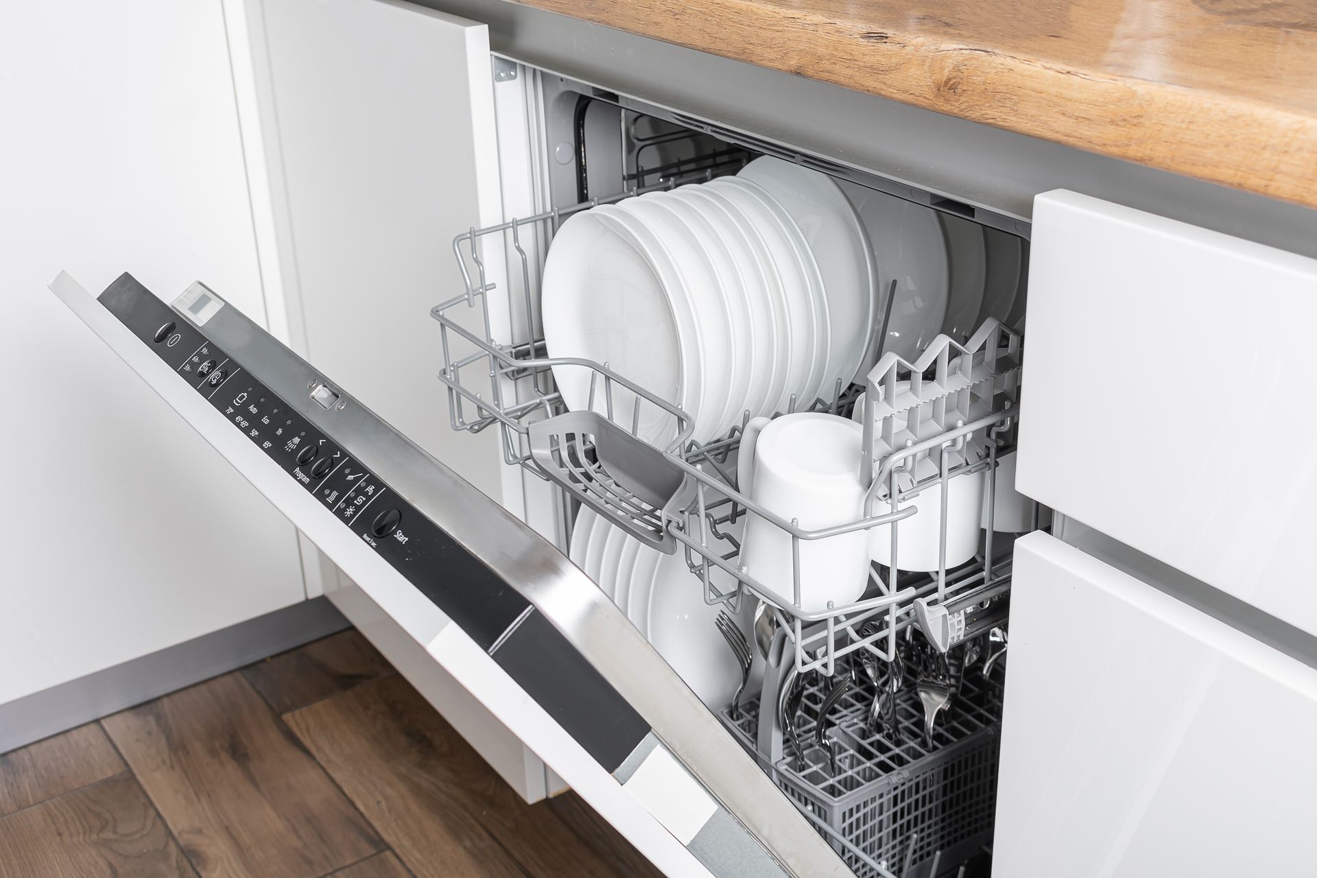Dishwasher with open door, loaded with white dishes and mugs in a white kitchen.