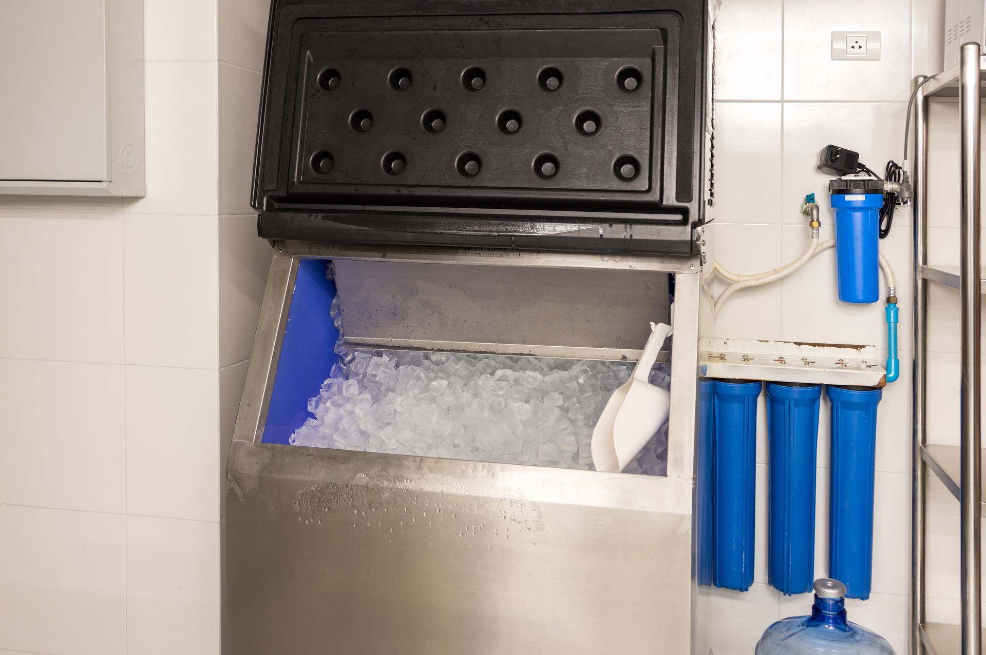 An open commercial ice machine filled with ice, a scoop, and a water filter system attached to the wall.