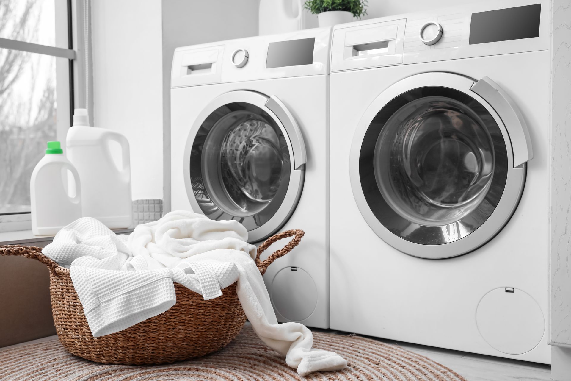 Two white washing machines beside a laundry basket of white towels; detergent bottles on a windowsill.