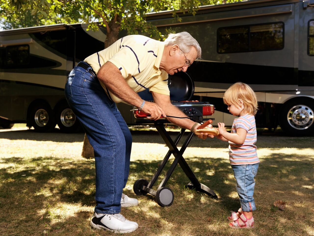 Grandpa living his ideal life while passing his Granddaughter a hotdog by the BBQ at a Campground
