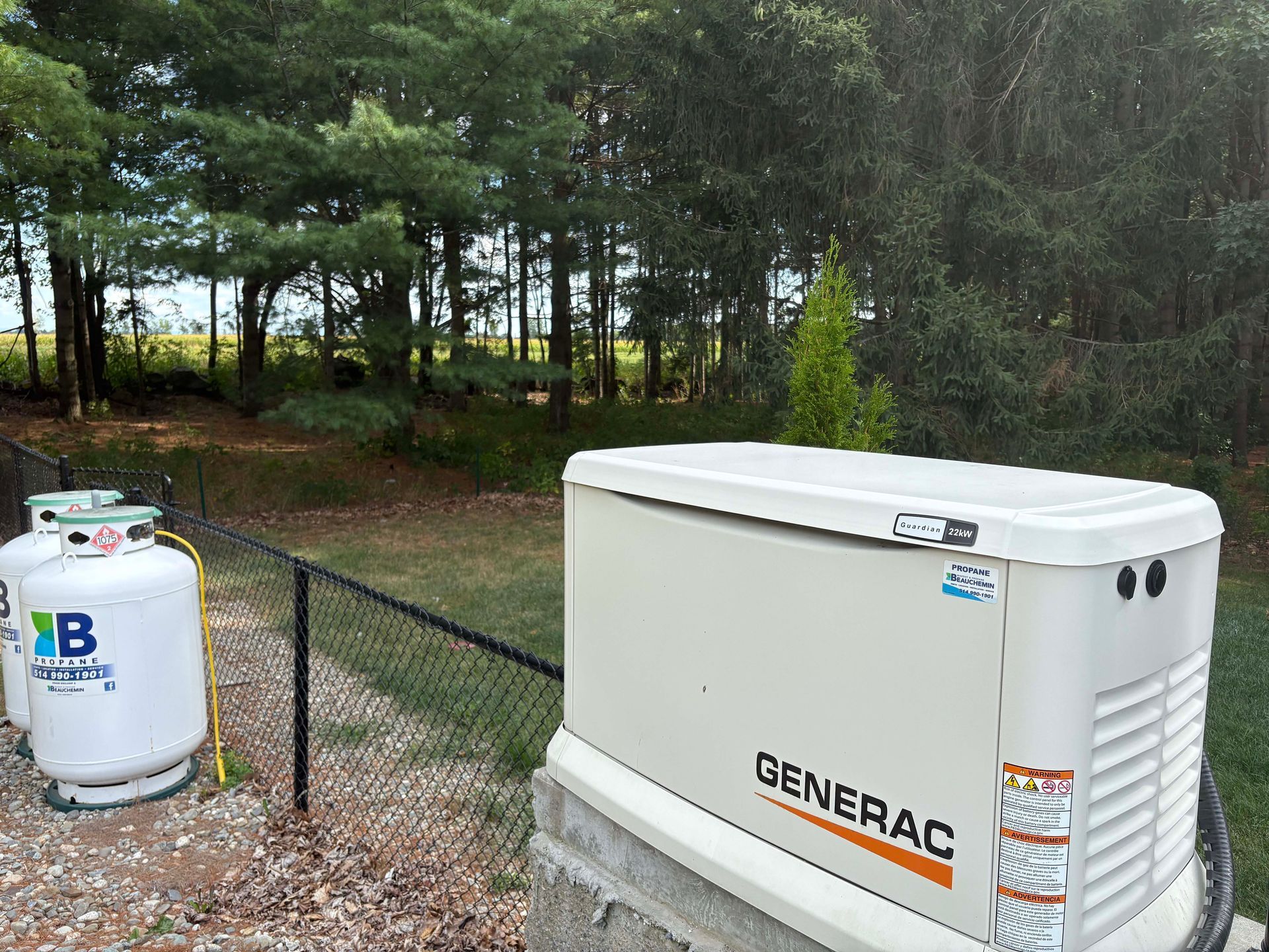 Un générateur Generac à côté de réservoirs de propane près d'une clôture noire et d'une zone boisée.