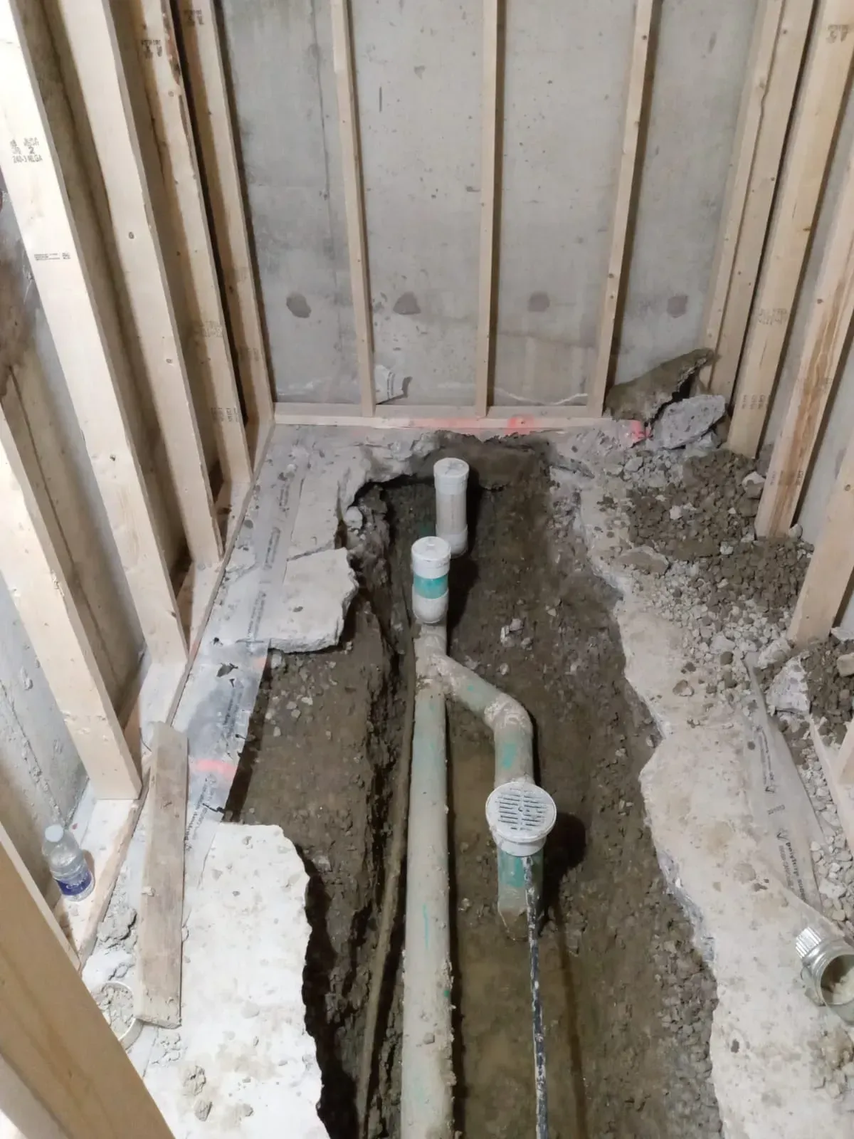Construction site with exposed green plumbing pipes and vertical wooden framing in a narrow, excavated area.