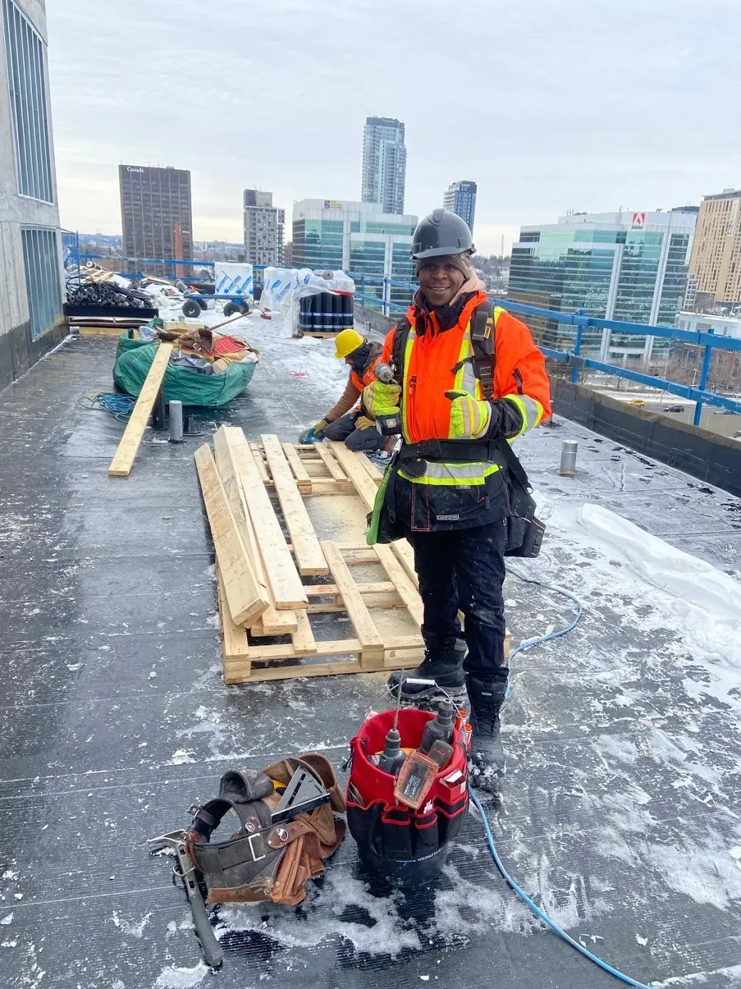 Construction worker on a rooftop wearing an orange vest and hard hat, smiling at the camera with a city skyline background.