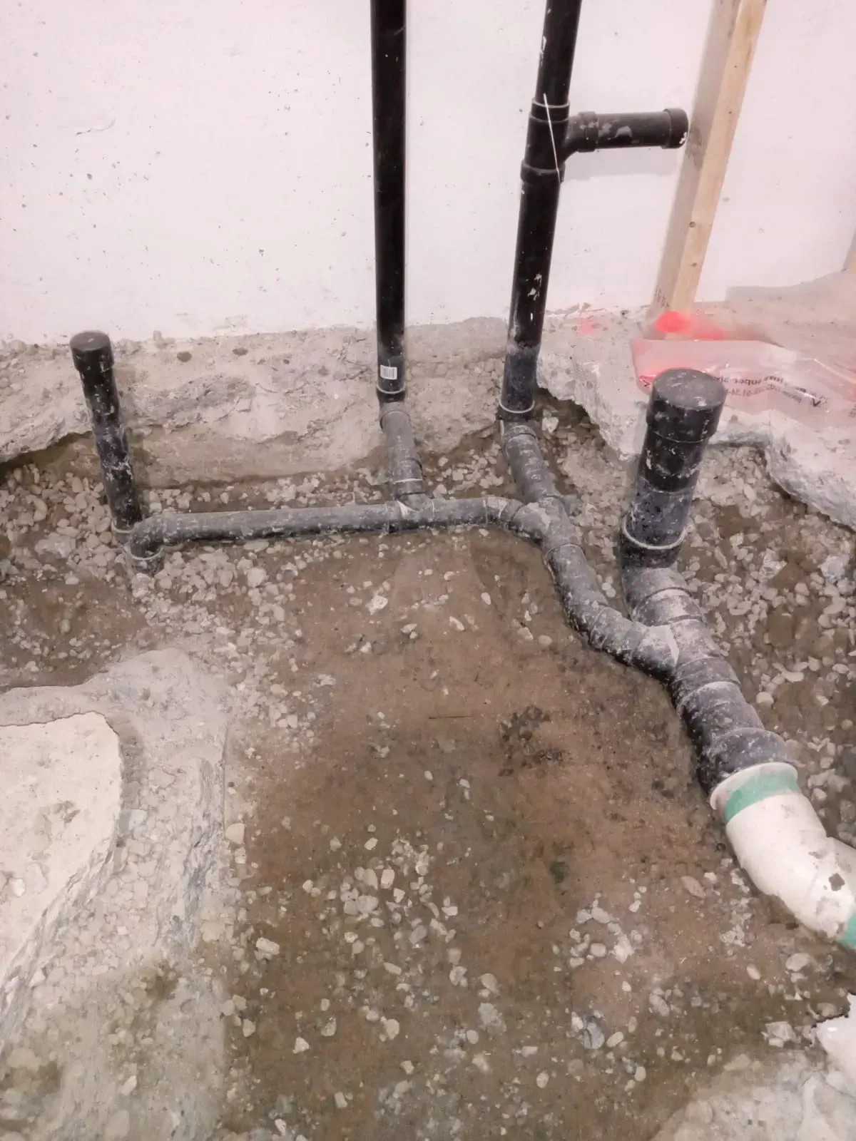 Plumbing pipes, black and gray, emerge from a concrete floor, indicating a bathroom installation in a construction setting.