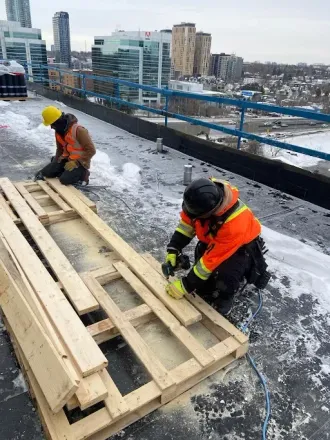 Two construction workers on a rooftop cut and assemble wood pallets.