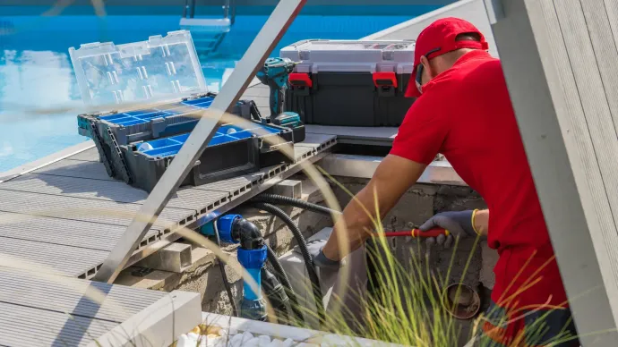 A pool technician in a red shirt and cap works on pool equipment, using tools, by a pool.