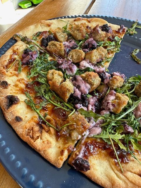 Pizza slice with sausage, arugula, and blue berry goat cheese on a blue plate.