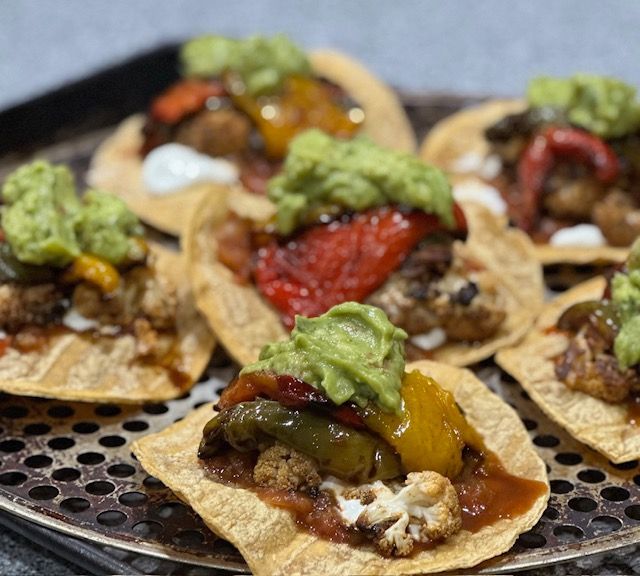 Six loaded tostadas on a perforated metal surface: roasted vegetables, guacamole, and a drizzle of sauce.