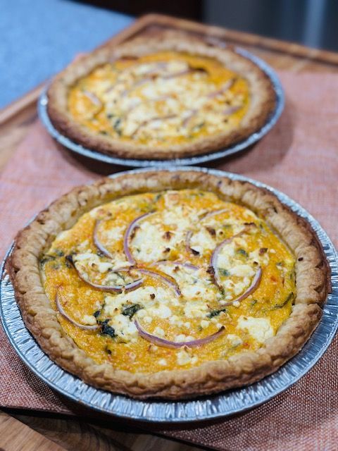 Two pies in foil tins, with golden crusts and toppings of red onion and white cheese.