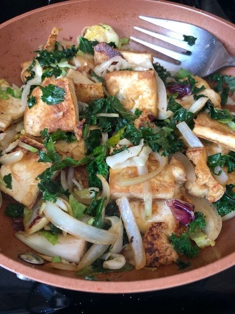 Tofu and vegetable stir-fry with kale, onions, and sprouts in a copper pan with a fork.