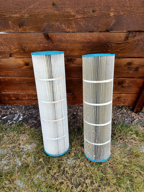 Two cylindrical pool filter cartridges with blue end caps standing upright on grass in front of a wood fence.