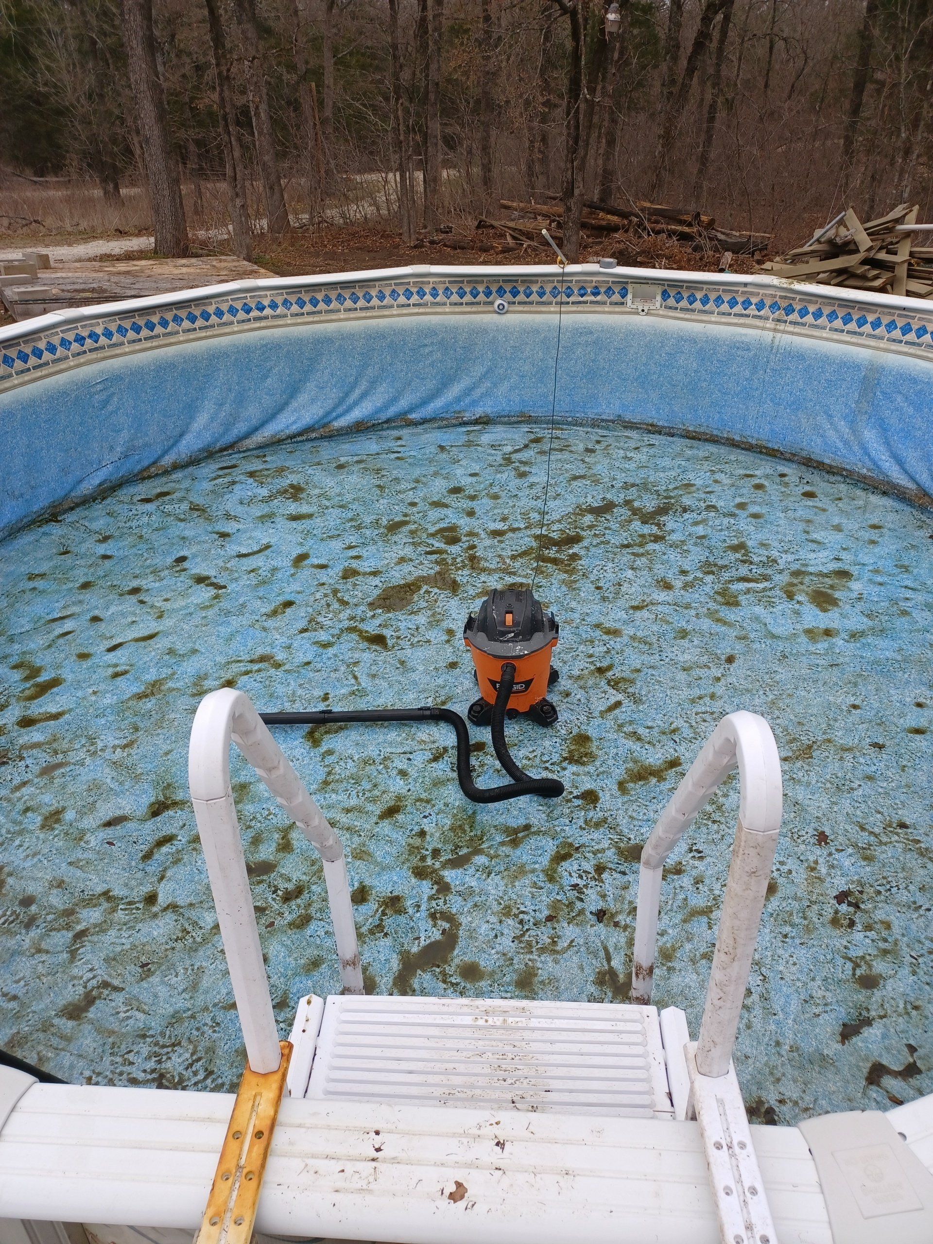 An orange shop vacuum sits in a dirty, empty above-ground swimming pool surrounded by woods.