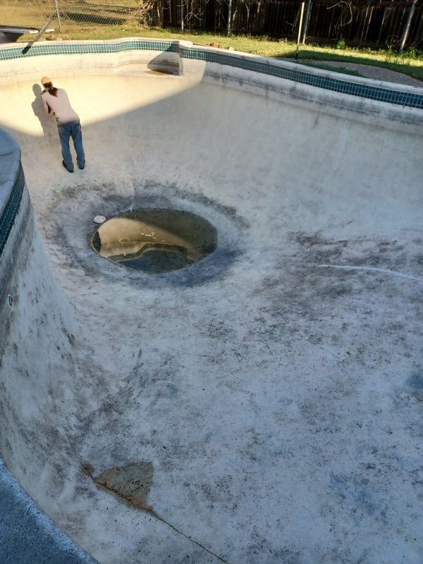 A person stands in a dry, empty swimming pool with a central deep end drain, showing signs of wear and surface staining.