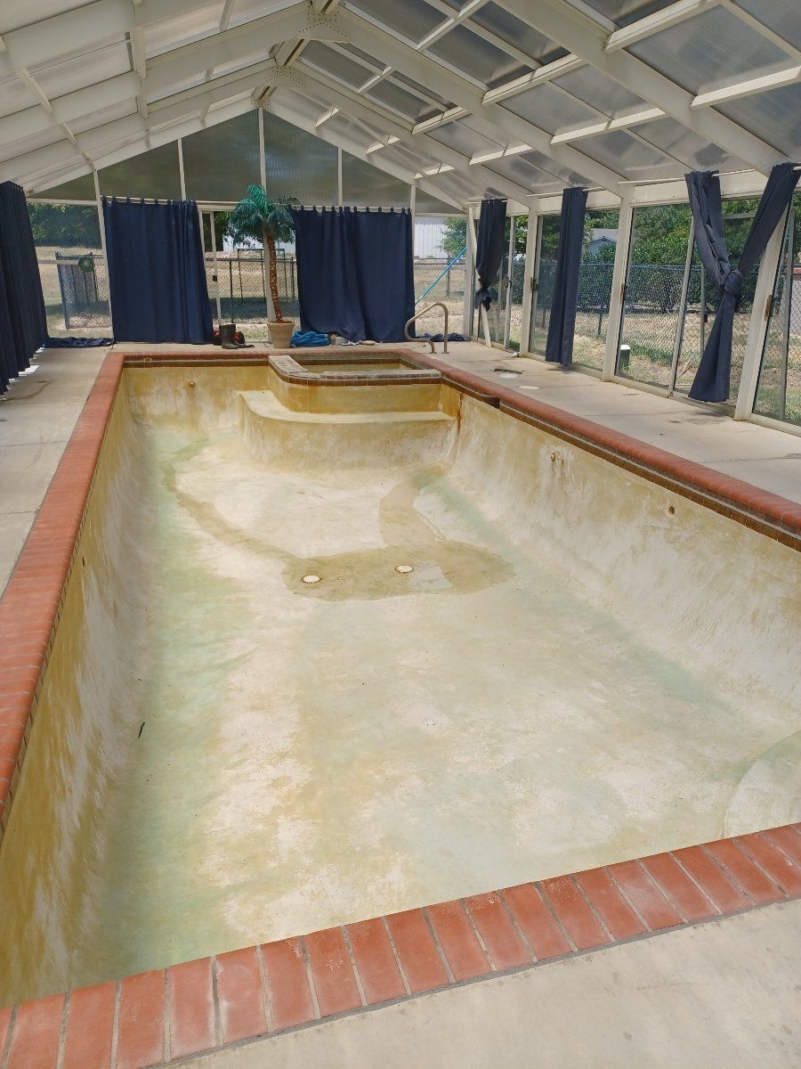An empty, stained swimming pool with a brick border, located inside a white-framed glass enclosure with dark curtains.