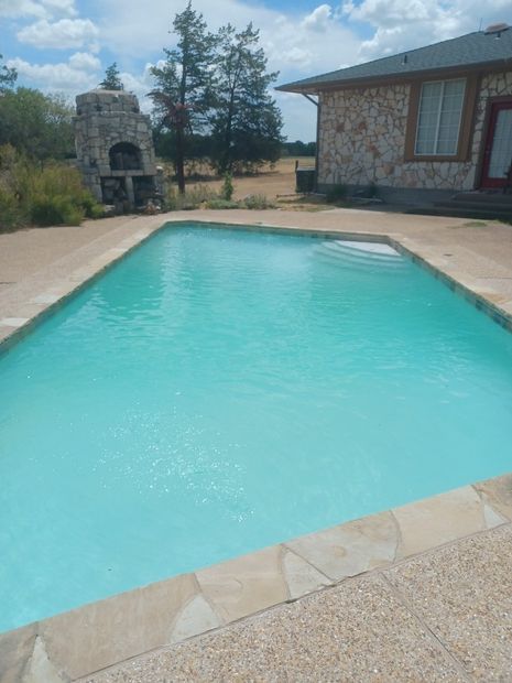 An outdoor rectangular swimming pool with turquoise water next to a stone house and a stone outdoor fireplace.