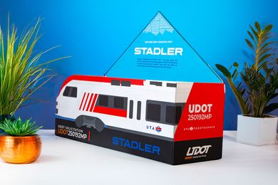 STADLER-branded white, red, and black train model on a tabletop with blue backdrop and plants