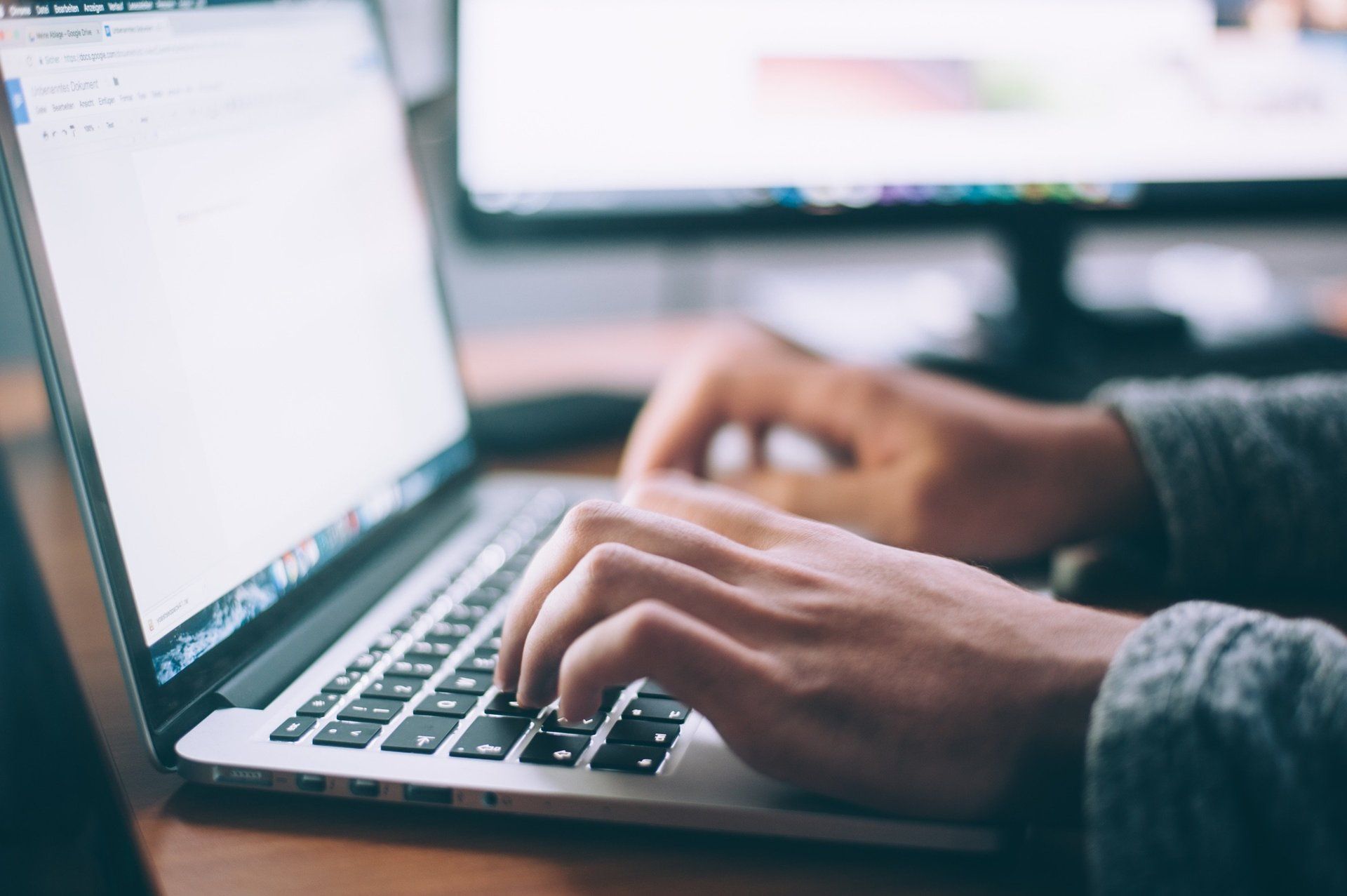 Hands typing on a laptop in a workspace with blurred monitors in the background