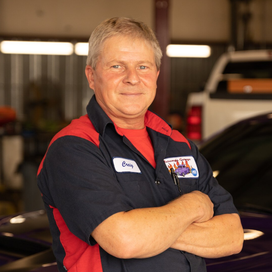 Mechanic, arms crossed, in a garage. Wearing work uniform with logo; red and blue. Smiling.