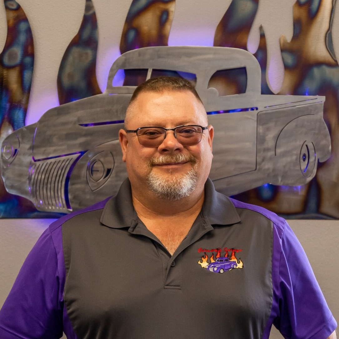 Man with glasses and a goatee in a gray and purple shirt stands in front of a metal truck sculpture.