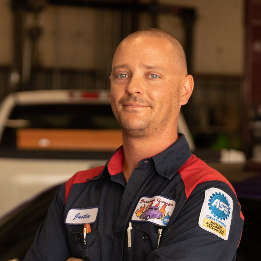 Man in mechanic uniform with folded arms, smiling. In auto shop setting.