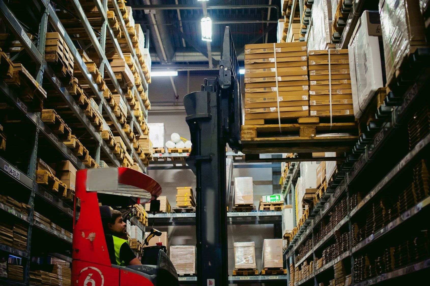 A forklift operator maneuvers a stack of boxed goods in a tall warehouse aisle lined with industrial storage shelves.